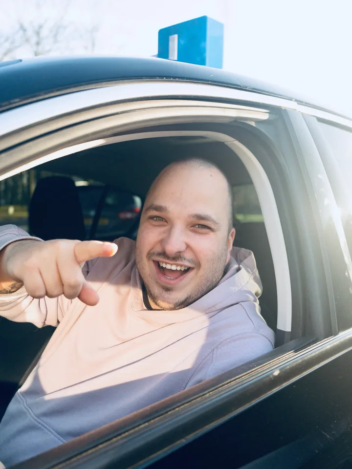 Bjorn van Lierop lacht achter het stuur van de lesauto en wijst naar de camera